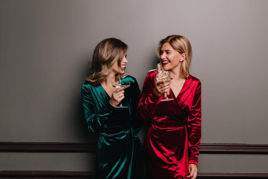 Two elegant escorts in red and green velvet dresses enjoying an intimate evening with champagne.