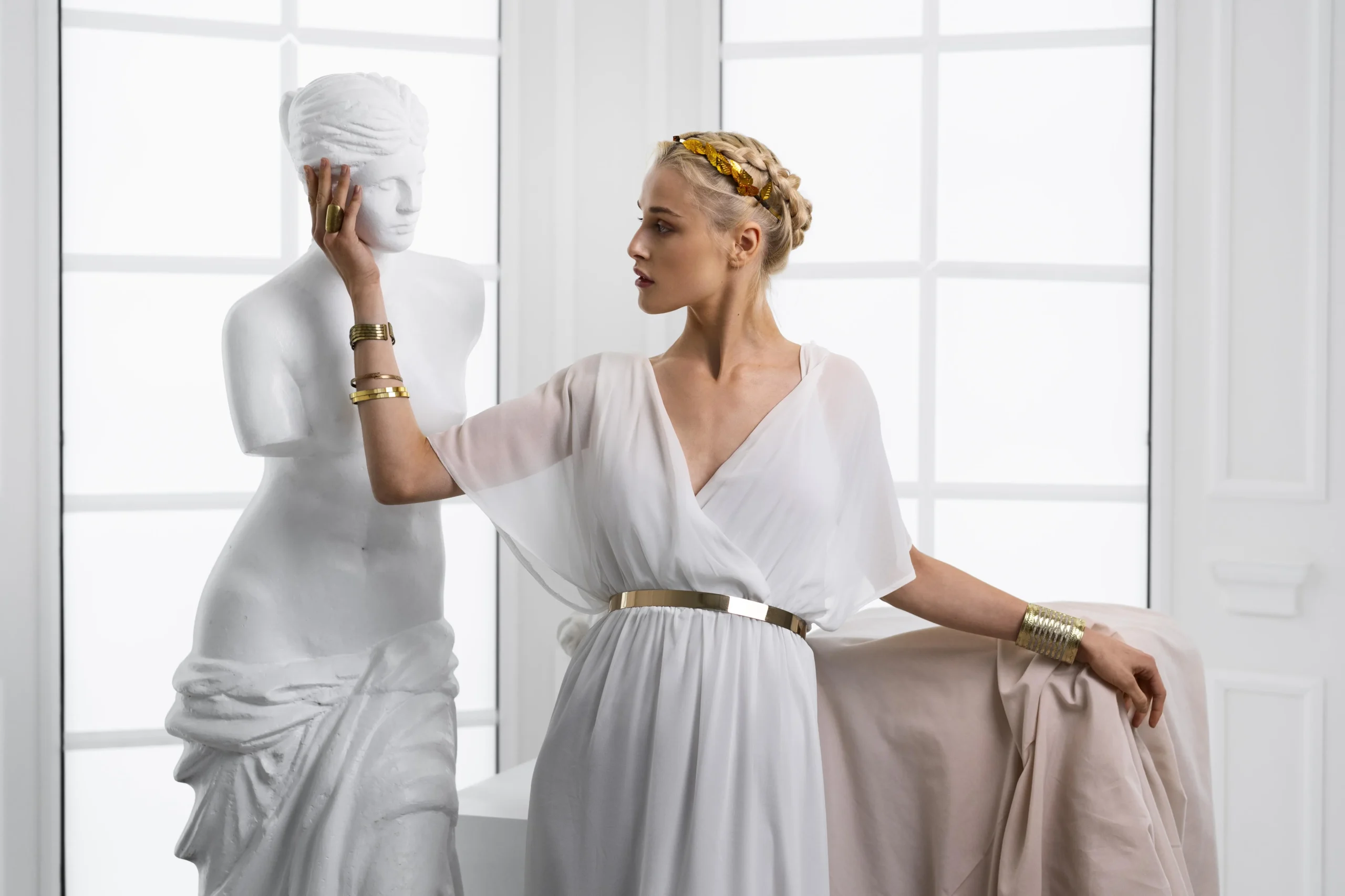 A graceful woman dressed in a flowing white gown with golden accessories posing beside a classical marble statue in a bright, elegant room.