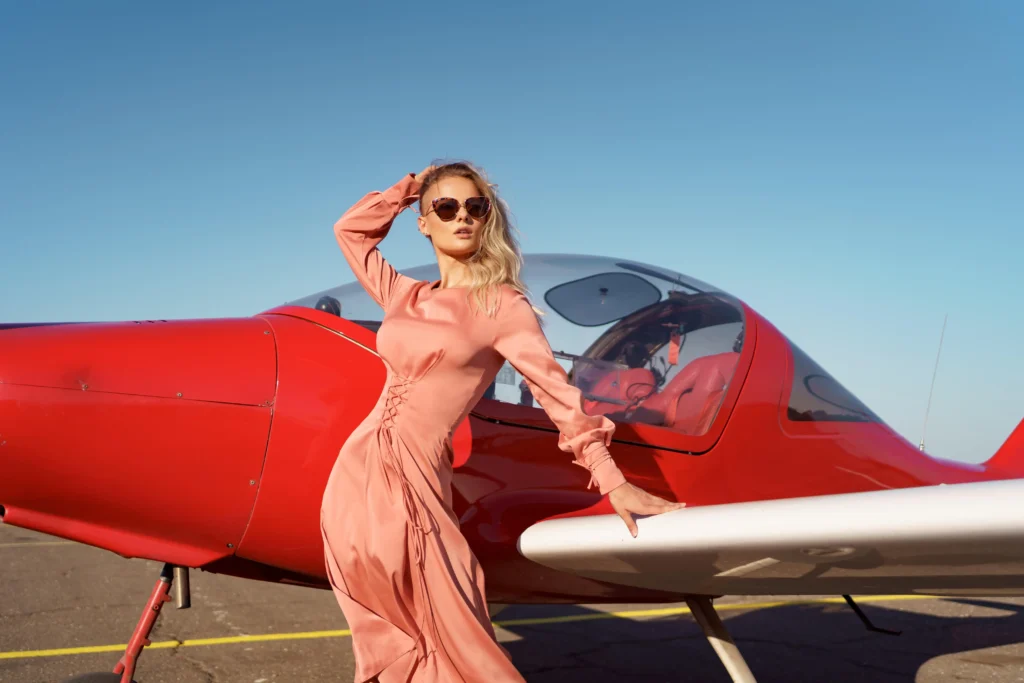 A stylish woman in a peach dress and sunglasses poses next to a red private jet under a bright blue sky, showcasing elegance and exclusivity.