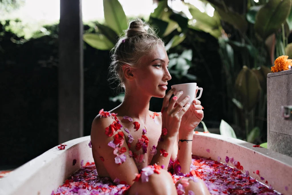 A peaceful woman soaking in a flower-filled bathtub outdoors, sipping tea and embracing a self-care ritual surrounded by lush greenery.